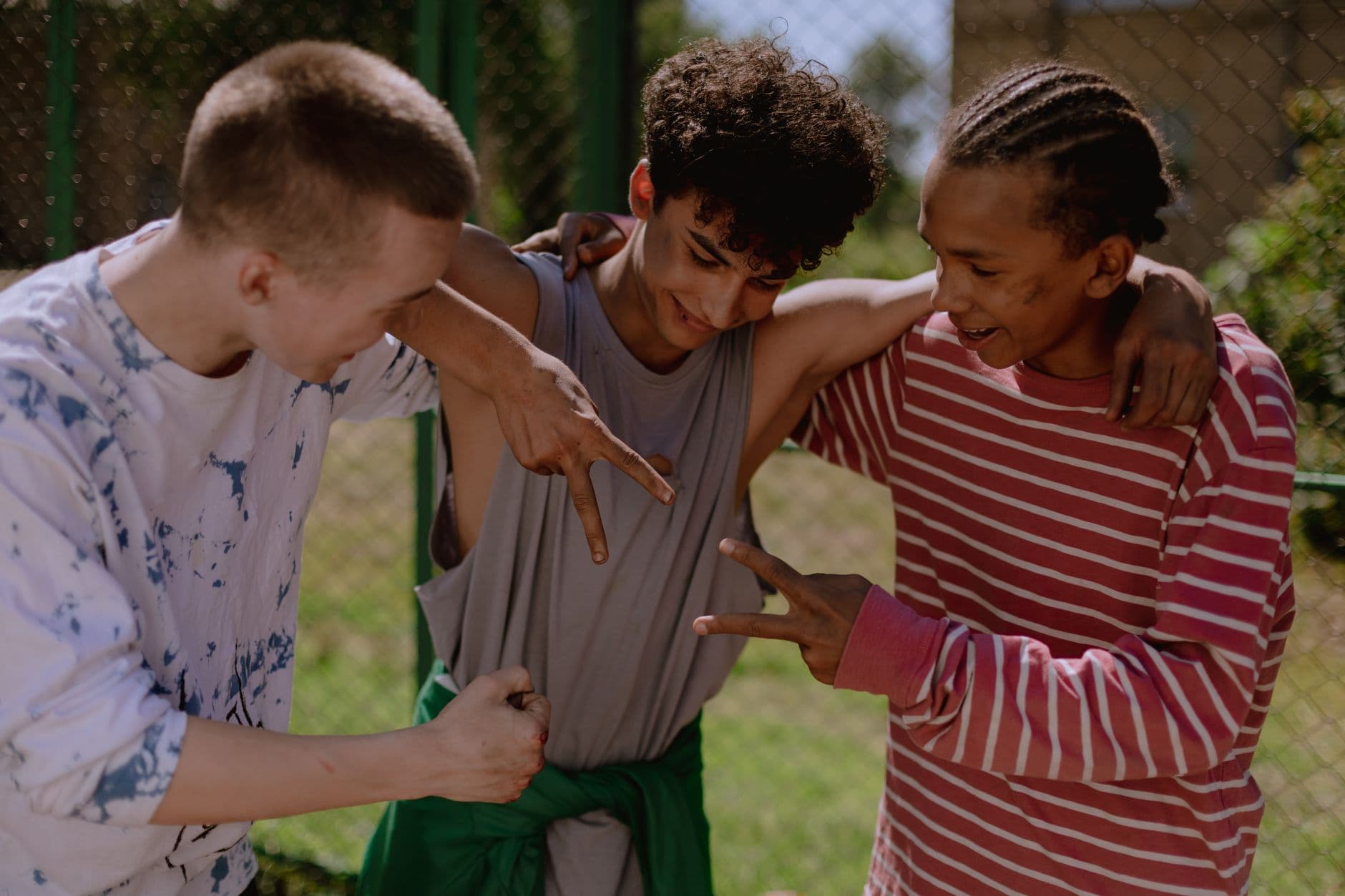 Three teenage friends playing rock, paper, scissors outdoors by a sports court, laughing with their arms around each other's shoulders.