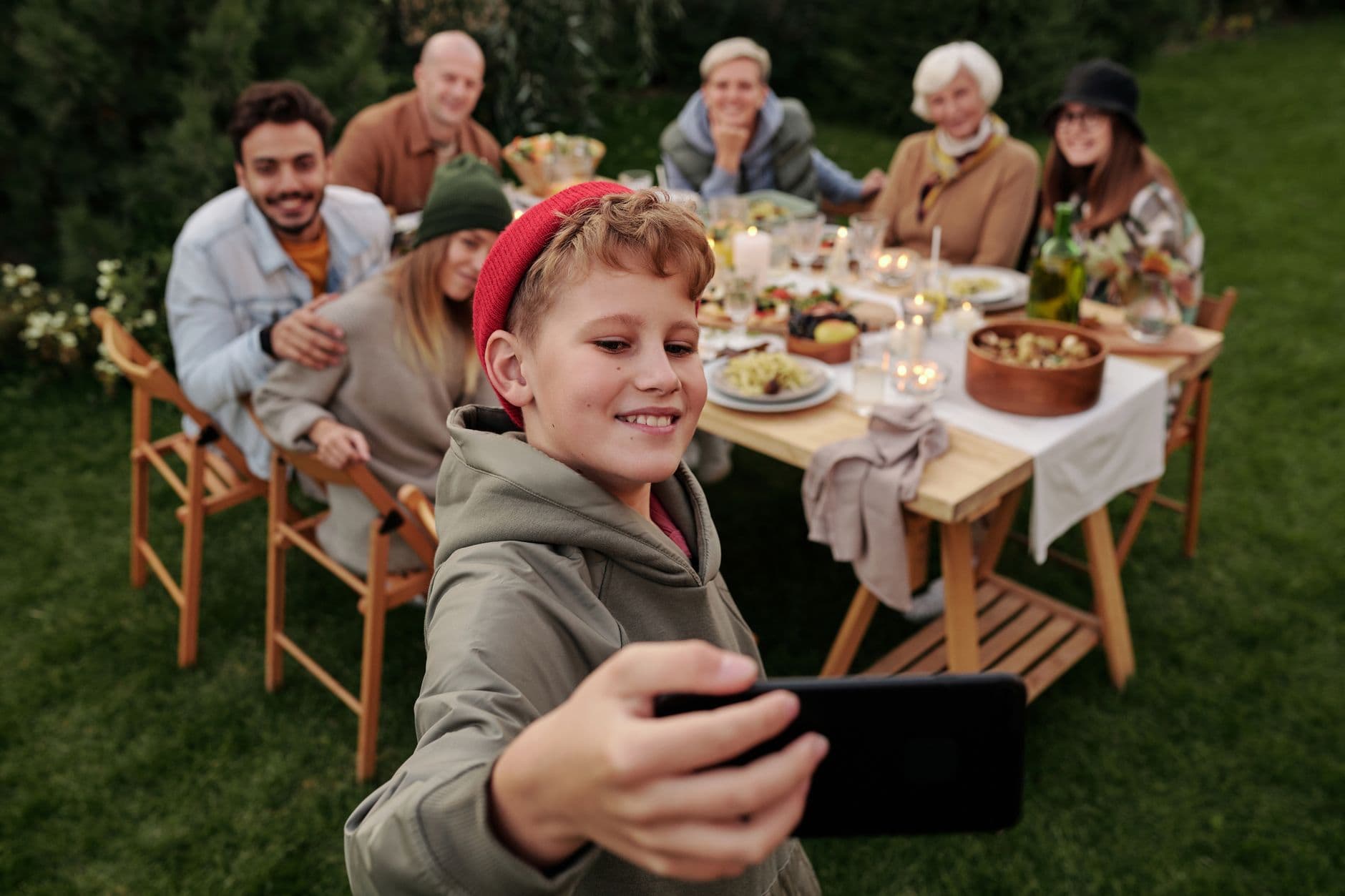 A boy takes a selfie in front of his family, who are sitting around a garden dinner table with food and candles.