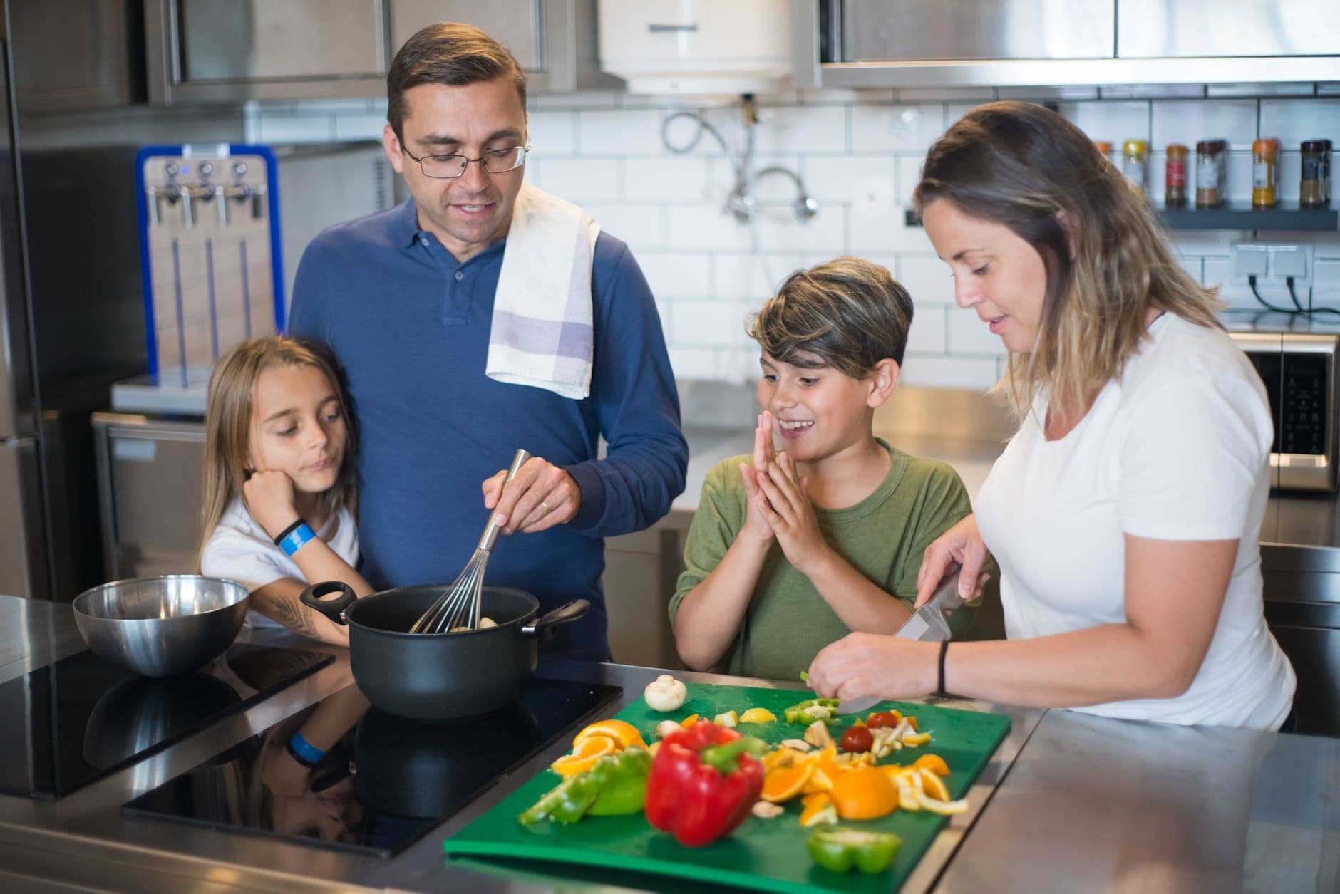 A family of four preparing a healthy meal together in a kitchen, chopping peppers and oranges on a green board.